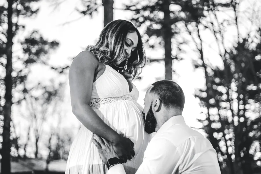 Black and white maternity portrait of a partner kissing an expectant mother's bump outdoors