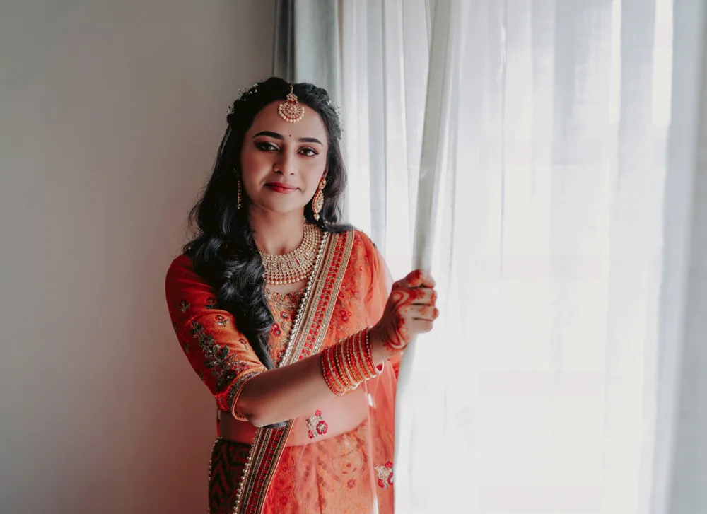 Bride in orange standing beside a window in soft daylight, bridal photography Bangalore